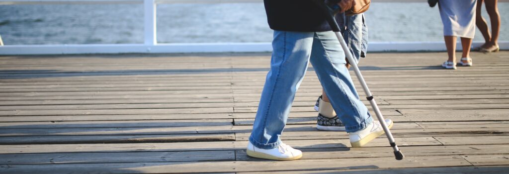 A close up image of a person walking on a boardwalk using a cane as a mobility aid