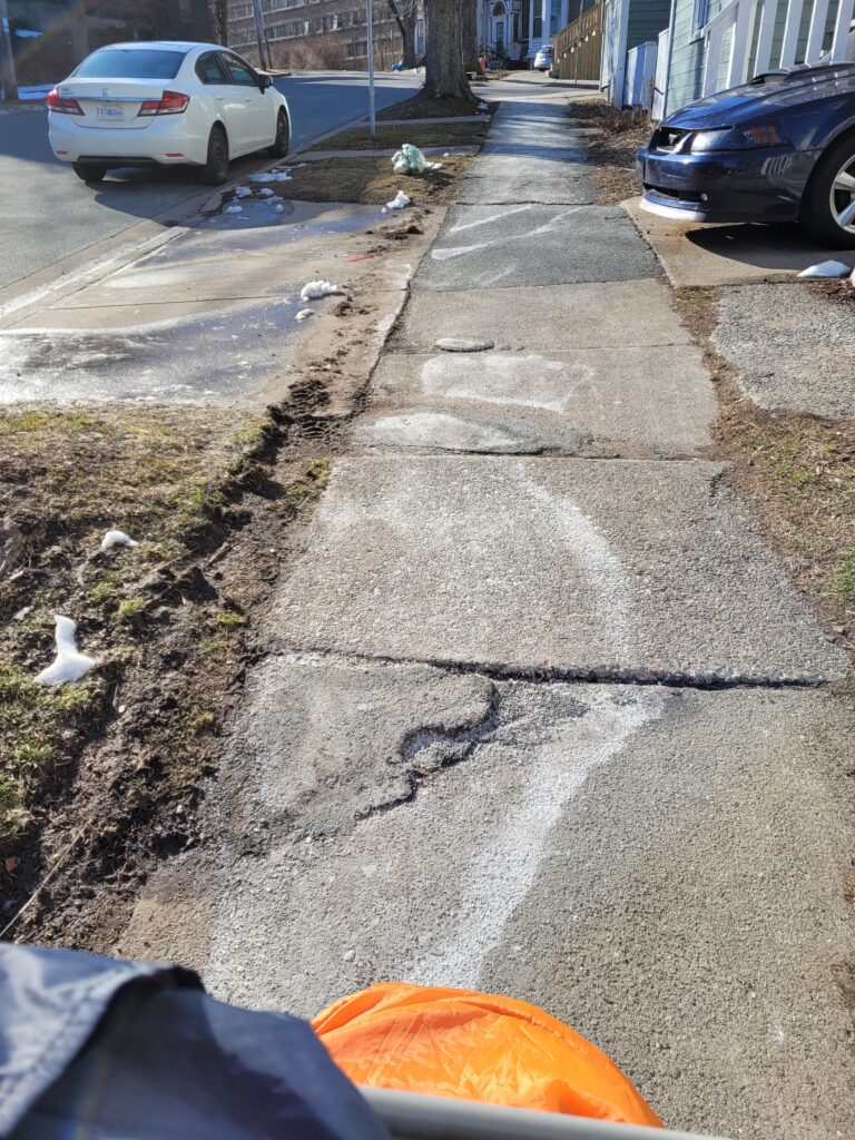 The sidewalk has many large cracks and raised edges between sidewalk slabs. The elevation changes abruptly along the sidewalk and there are salt marks showing where water has pooled. 