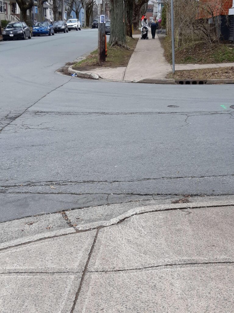 An unmarked intersection with curb cuts that do not align. Curb cuts direct diagonally into the intersection. A couple is in the distance pushing a stroller down the sidewalk.
