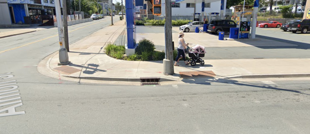 A street corner with wide sidewalks and two unidirectional curb cuts each with a tactile attention indicator. A woman is pushing a stroller along the sidewalk.