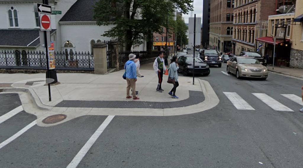 A bump out with tactile attention indicator wrapped around the edge of both sides of the corner for crossing. There are bright white painted lines for each crossing direction and a group of people are approaching to cross.