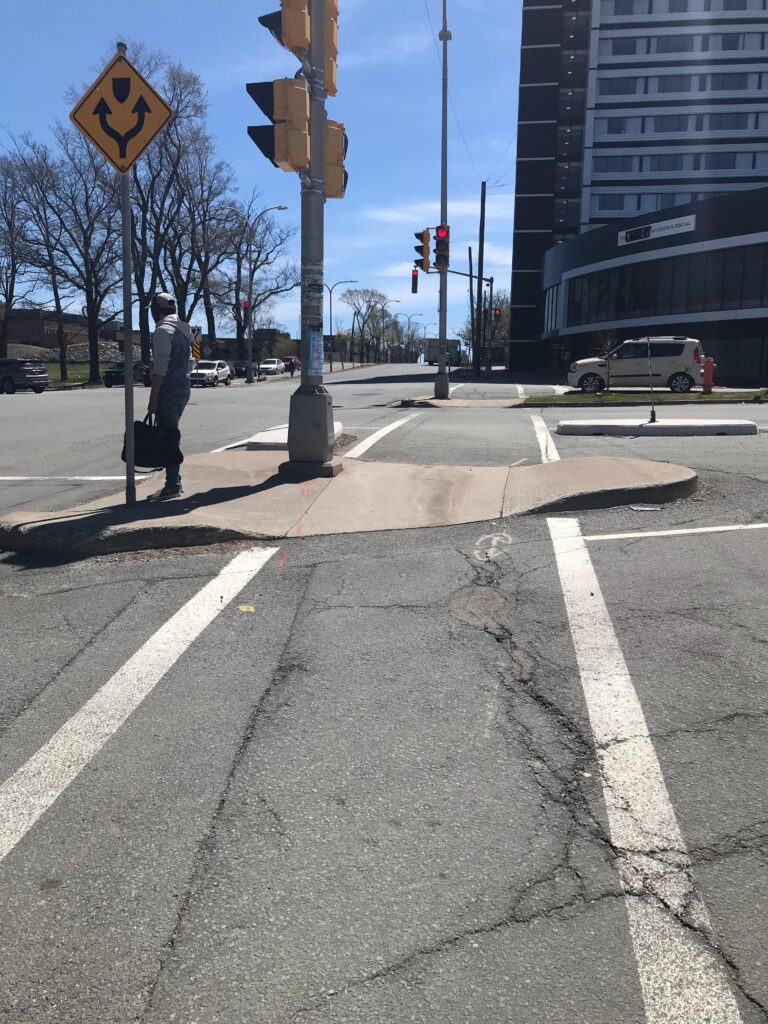 The crossing at Quinpool Rd is shown straight-ahead, with two traffic islands in the path of crossing. Both are graded but the curb cuts are somewhat narrower and misaligned from the painted crossing lines. White painted lines appear bright, straight, and continuous across the street. There are some cracks in the pavement and no tactile attention indicators present.