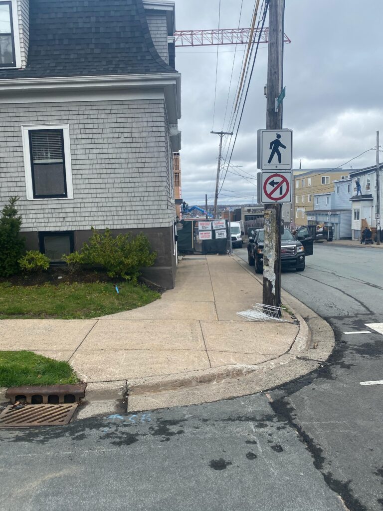 A street corner with a very narrow curb cut and wire litter on the sidewalk. There is construction down the street that blocks the sidewalk without any signage or an alternate path of travel around the obstacle. 