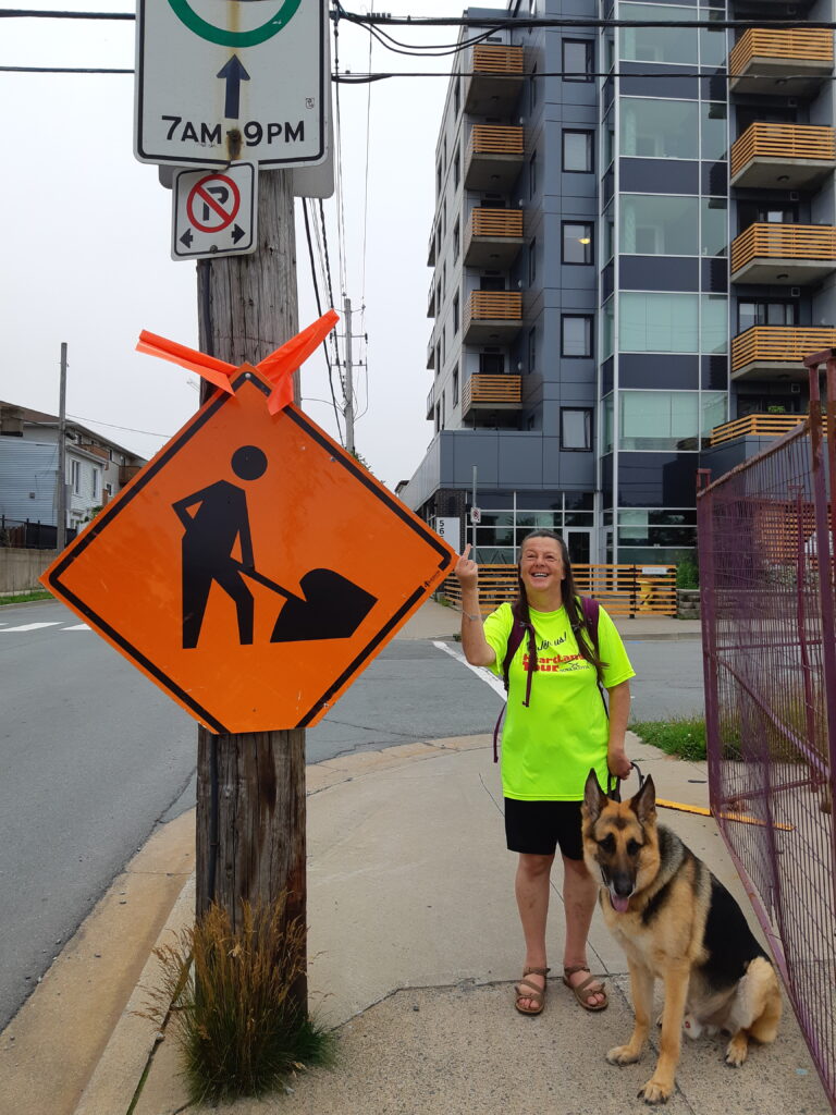 A woman stands on a sidewalk giving the finger to a diamond shaped construction sign strapped to a telephone pole at head level, with her guide dog sitting next to her.
