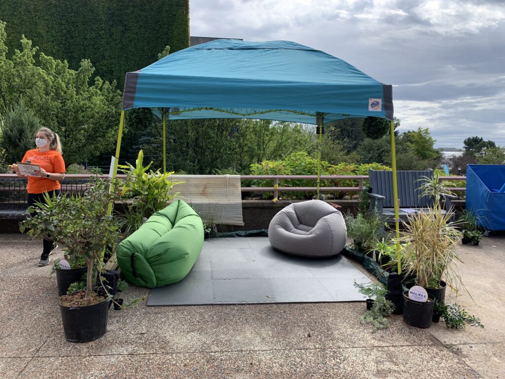 A temporary blue tent canopy is set up with inflatable seating and soft tile flooring underneath, surrounded by potted plants.