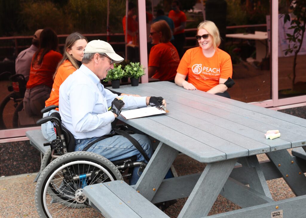 A man using a wheelchair sits at an accessible picnic table with two researchers wearing orange PEACH T-shirts.