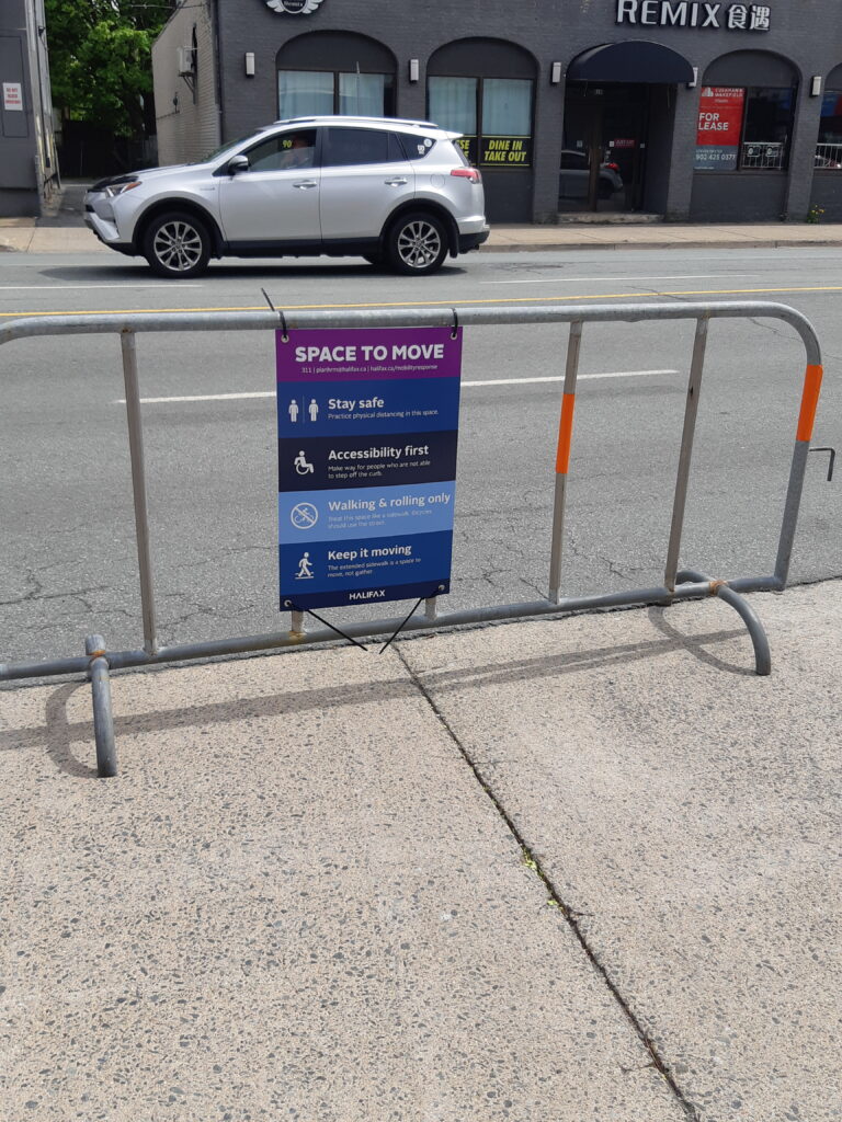 A metal barrier is placed in street parking to extend the sidewalk for pedestrian movement. A sign is posted on the barrier describing HRM's Space to Move program during the pandemic.