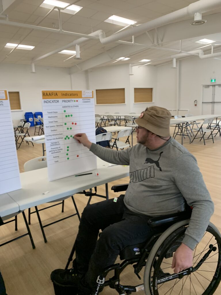 A man using a wheelchair and wearing a tan hat reaching out to place a sticker on a poster beside him.