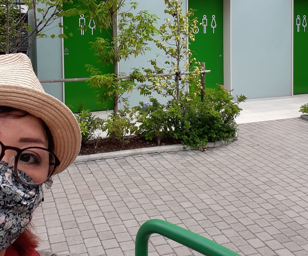 A woman wearing a hat, glasses, and a face mask poses in front of a row of green public washroom doors.