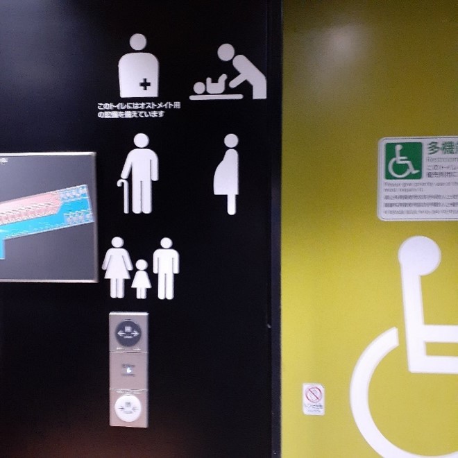 Large white icons on a black wall indicating a change table, family washrooms, older adult-friendly, and pregnant person-friendly, next to a yellow door with an extra large universal symbol of accessibility.
