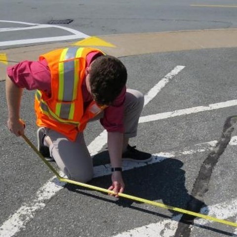 A man wearing a reflective vest crouching down to measure markings in a parking lot. 