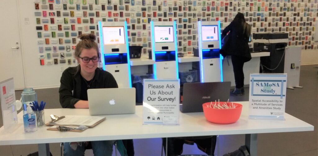 A smiling girl sits at a kiosk in a library advertising the samosa survey