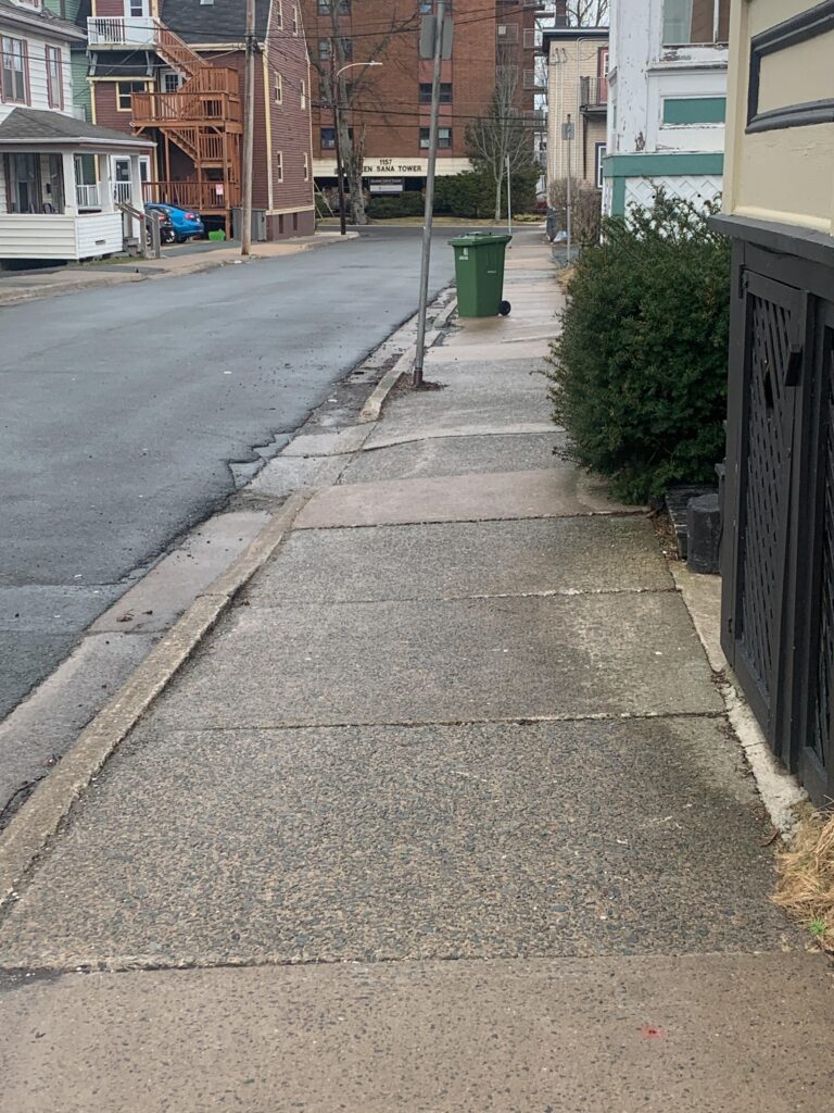 A concrete sidewalk stretches out ahead with many bumps in its elevation. A hedge is encroaching onto the sidewalk, and a fixed parking sign and green bin obstruct the sidewalk.