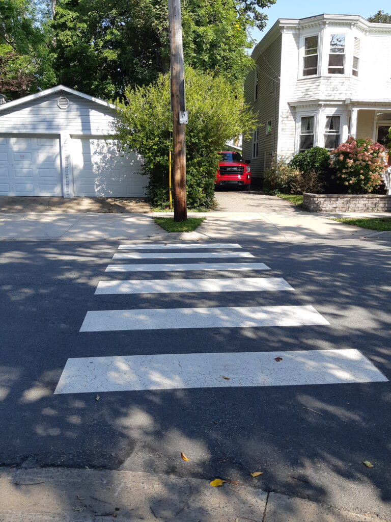 Bright white zebra lines painted at a three-way street intersection lead to a raised curb where there is a grassy boulevard and telephone poll with driveways to either side.