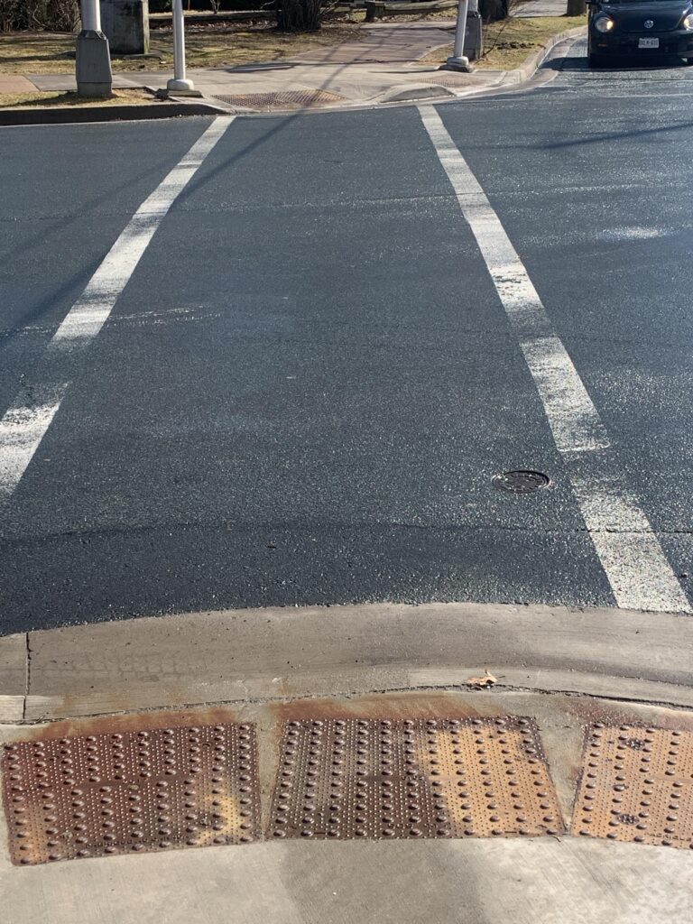 A crossing with straight painted line lead across a street with curb cuts on either end. Both curb cuts have tactile attention indicators. 