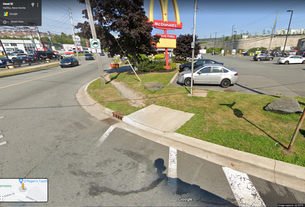 Crosswalk leads to a single sidewalk square on a grass island between the road and a parking lot.