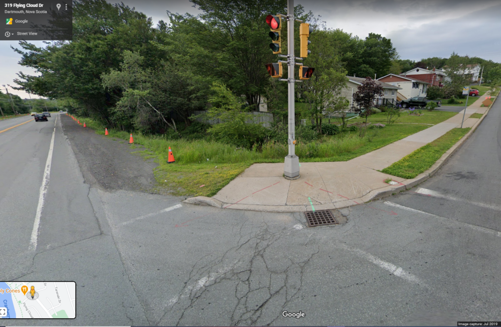 One sidewalk leads to a street corner with a traffic light located in the path of pedestrian travel. The sloping curb cut appears to be steep and leads to a grate where the pavement meets the curb cut.