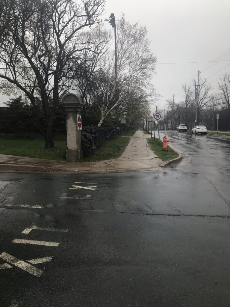 Concrete curb cut is cracked at the entrance to Alumni Cres. It appears to have just rained and all surfaces are slick.