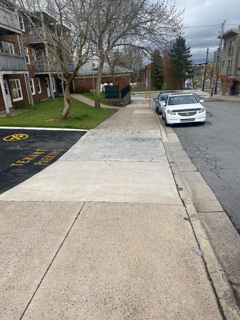 Wide concrete sidewalk that is cut for driveway access. Cars are parked along the street.