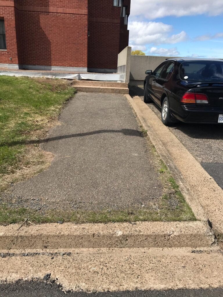 A paved approach to a building entrance is obstructed by a curb and a set of three steps.