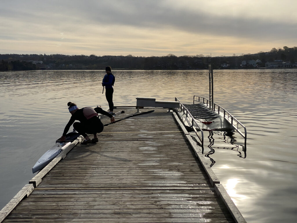 Two people stand on a dock overlooking a lake. There is a structure with railings fastened along the dock and into the water.