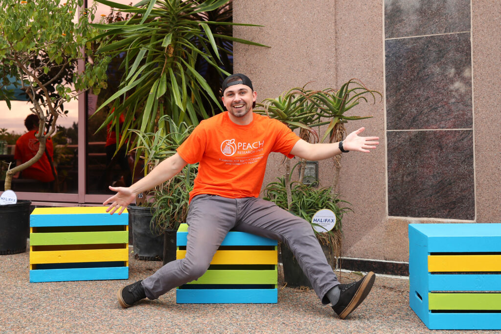 A young man wearing an orange PEACH Research T-shirt smiles and poses with his arms wide to show off colourful crate seats built for the event