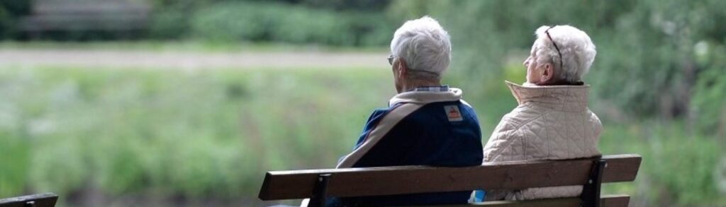 Two adults with grey hair seated on a bench overlooking a green landscape.