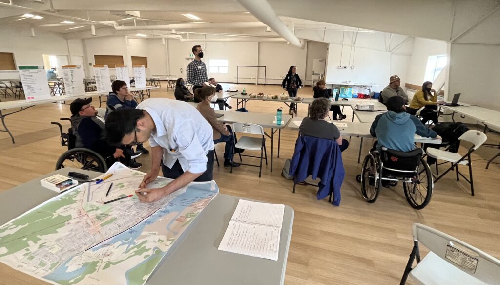 A young man leaning over a map on a table with people engaged in a roundtable discussion in the background.