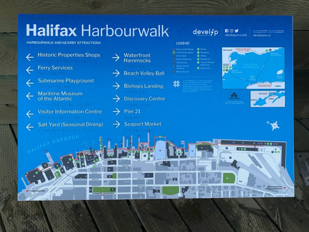 A blue sign showing lists of destinations in white text and braille with a map of the Halifax boardwalk along the bottom.