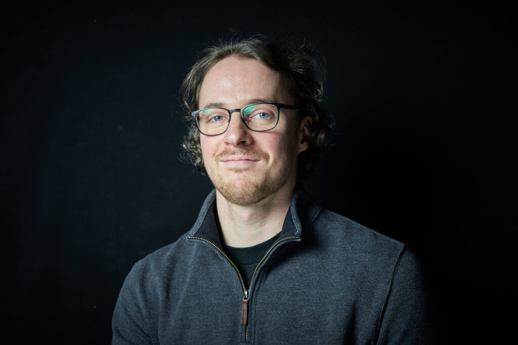 A smiling young man wearing a dark blue sweater and glasses.