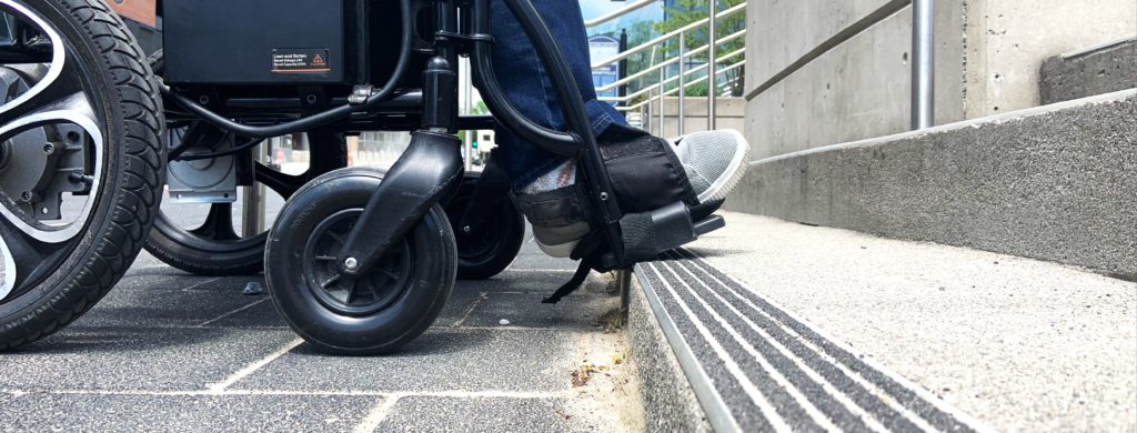 A foot of a person using a wheelchair encountering a step at the bottom of a staircase.