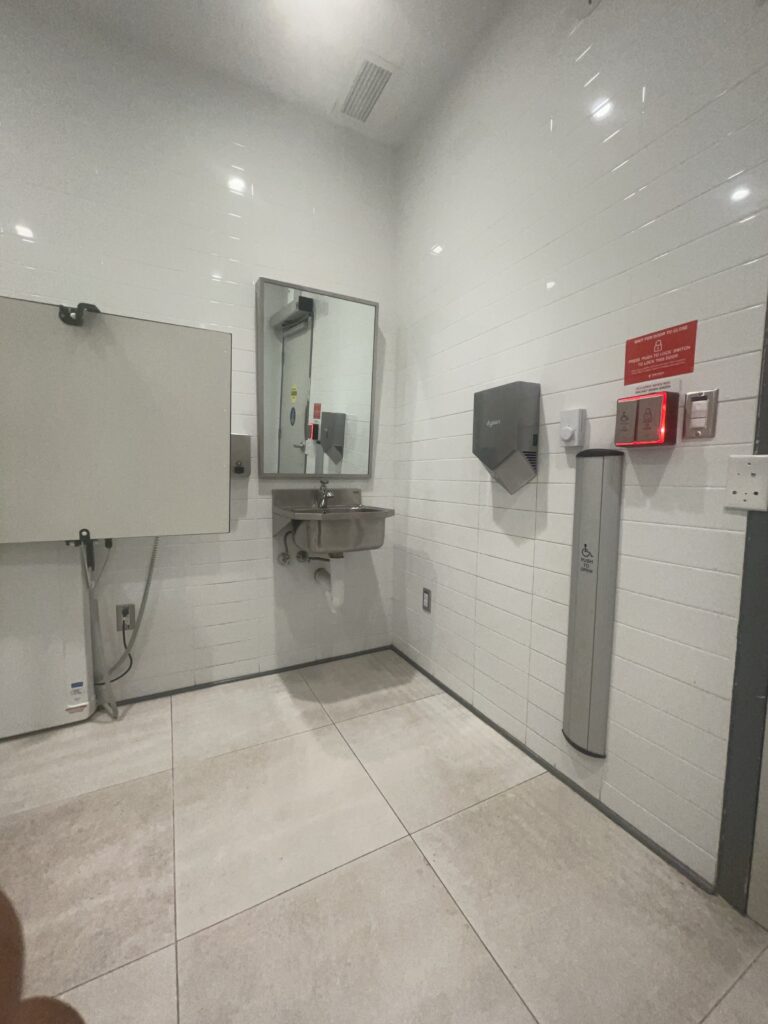 Bathroom interior with wall-mounted sink with leg room underneath, an adult-sized change table, and pushbuttons to open, close, lock, and unlock the door.
